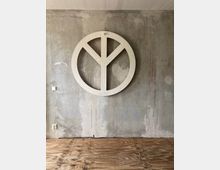 Eine Betonwand mit sichtbarer Struktur, vor der ein großes, weißes Peace-Zeichen angebracht ist. Der Boden besteht aus unbehandelten Holzplatten, und an der Wand befindet sich eine Steckdose sowie ein Lichtschalter.