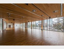 Großer, moderner Veranstaltungsraum mit Holzfußboden und einer Decke aus Holzpaneelen mit eingebauten Rundlüftern. Die Wände bestehen größtenteils aus bodentiefen Glasfenstern, die den Blick auf urbane Gebäude im Hintergrund ermöglichen.