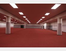 Weitläufiger, rechteckiger Innenraum mit rotem Teppichboden, weißen Wänden und zahlreichen runden Säulen. Die Decke ist mit rechteckigen Deckeneinbauleuchten ausgestattet, die eine gleichmäßige Beleuchtung erzeugen.