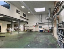 Große, funktionale Werkhalle mit grünem Betonboden, weiß gestrichenen Wänden und einer Zwischendecke. An der rechten Seite befinden sich zahlreiche Lüftungsrohre und Regale mit Farben oder Materialien, während links Arbeitsbereiche und Regale zu sehen sind.