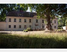 Zweistöckiges weißes Herrenhaus mit symmetrischer Fassade, vielen Fenstern und einem gewölbten Eingangsbereich, umgeben von Bäumen und einem naturbelassenen Hof mit hohem Gras.