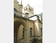 Helle Fassade eines historischen Gebäudes mit beige-gelben Wänden, Rundbogenfenstern und dekorativen Türmen. Im Vordergrund befinden sich arkadenartige Bögen und ein schmiedeeisernes Geländer.