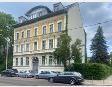 Straßenszene mit einem historischen mehrstöckigen Wohngebäude im Stil des 19. Jahrhunderts, mit gelber Klinkerfassade, verzierten weißen Fensterrahmen und einem gewölbten Dachgeschoss. Vor dem Gebäude befinden sich parkende Autos, Bäume und eine Hecke.