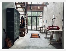 Großer Werkstatt- oder Atelierraum mit Betonboden, Holztreppe und großen Glastüren, die auf einen begrünten Außenbereich führen. Im Raum stehen Arbeitsbänke und Tische, daneben ein großes dunkelgrünes Metallschrank-Element, ein kleiner Teppich und eine an die Wand gelehnte Gitarre.