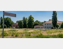 Ein Wegweiser mit der Aufschrift „Bahnhof“ und einem Fahrrad-Symbol steht vor einer Wiese. Im Hintergrund sind mehrere helle Gebäude mit roten Ziegeldächern und Bäume zu sehen.