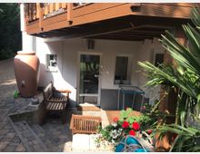 Unterer Bereich eines Wohnhauses mit gepflasterter Terrasse, Holzbank, großer Terrakotta-Wasseramphore und blühenden Pflanzen in Töpfen. Ein Holzgeländer und überdachte Elemente markieren den Übergang zum Obergeschoss.