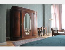 Schlafzimmer mit retro-stilvoller Einrichtung, bestehend aus einem großen Holzkleiderschrank mit ovalem Spiegel, einem kleinen Schreibtisch und einem Stuhl. Der Raum hat eine gemusterte Tapete, Holzboden, eine Stehlampe und helle Vorhänge, die Tageslicht hereinlassen.