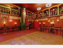 Innenansicht eines Theatersaals im Jugendstil mit roten Wänden, goldenen Stuckverzierungen und einer geschwungenen Wendeltreppe. Oben gibt es eine Galerie mit Tischen und Stühlen, und unten stehen Tische mit Lampen und Stühlen auf einem Holzboden.