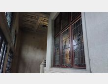 Innenansicht eines historischen Gebäudes mit hellem Steinmauerwerk, großen Fensterflächen aus Buntglas mit floralem Muster und Holzeinfassungen. Deckenverzierungen und ein dekoratives Säulenelement ergänzen die Gestaltung.