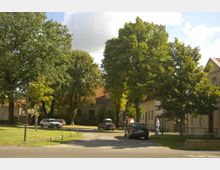 Straßenszene mit Bäumen und parkenden Autos vor mehreren Wohnhäusern mit roten Ziegeldächern. Der Bereich ist von Grünflächen und breiten Bäumen umgeben, unter einem hellen Himmel mit vereinzelten Wolken.
