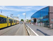 Straßenszene in einer Stadt mit einer gelben Straßenbahn auf der linken Seite, einem modernen Gebäude mit blauer Glasfassade und Geschäften auf der rechten Seite, sowie einem Gehweg mit Fußgängern und abgestellten Fahrrädern. Im Hintergrund sind ein weißes Gebäude und grüne Bäume sichtbar.