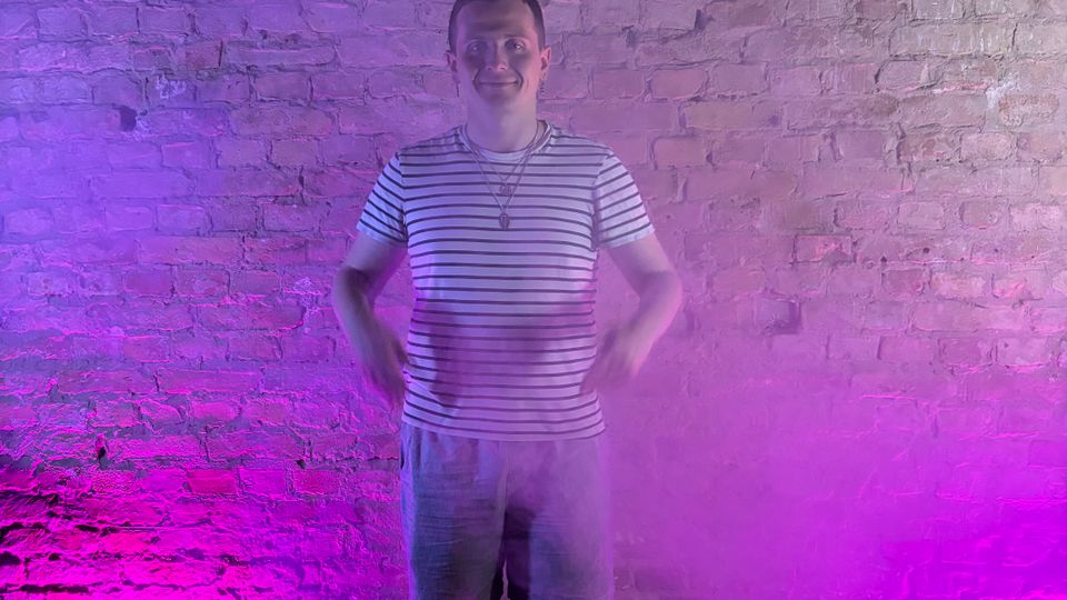 Indoor studio space with an exposed brick wall backdrop, lit by purple and pink uplights near the floor. Dark wood flooring and light haze create a soft, smoky effect across the lower part of the scene.