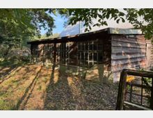 Holzgebäude mit braunen Brettern und großen, rechteckigen Fenstern, die teilweise beschädigt sind, umgeben von Laub und einer bewaldeten Umgebung. Das Dach besteht aus Wellblech, und der Boden ist mit Herbstlaub bedeckt.