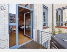 Blick auf einen Balkon mit Holzfußboden, einem Grill und Glasgeländer, der an ein Gebäude mit weißen Ziegeln und blauen Fensterrahmen grenzt. Durch eine große geöffnete Glastür ist ein Essbereich mit Backsteinwänden und modernen Stühlen sichtbar.