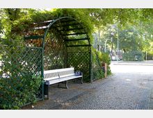Überdachte Sitzbank mit weißer Holzbank innerhalb einer Metallpergola mit Rankgittern, bewachsen mit grünem Laub. Der Bereich befindet sich an einer gepflasterten Fläche in einer städtischen Umgebung mit Bäumen und Laternen im Hintergrund.