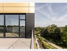 Moderne Gebäudeansicht mit großen Glasfenstern und goldener Fassadenverkleidung auf der linken Seite, daneben eine Aussicht auf einen grünen Park mit einem geschwungenen Spazierweg bei klarem Himmel. Der Vordergrund zeigt eine helle Holzterrasse.