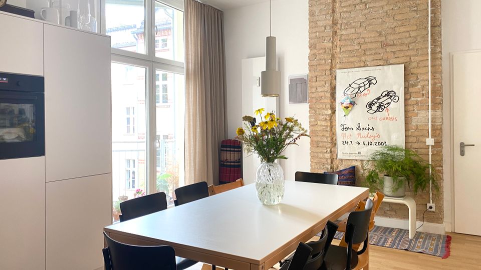 Heller Ess- und Küchenbereich mit großem rechteckigem Esstisch und mehreren schwarzen Stühlen, neben hohen Fensterfronten mit Vorhängen. An der Wand befindet sich eine freigelegte Ziegelwand mit Poster, Pflanzen und Hängeleuchte; links sind weiße Einbauschränke mit Backofen.