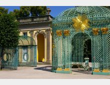 Dekoratives Gartenpavillon mit grüner Metallstruktur und kunstvollen Gittern, verziert mit goldenen Ornamenten und einer goldenen Sonnenfigur an der Spitze. Im Hintergrund gelbes Gebäude mit klassizistischen Säulen und einem Rundbogen, umgeben von Bäumen.