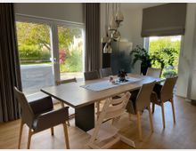 Esszimmer mit einem großen Holztisch und acht beigefarbenen Stühlen, beleuchtet durch eine Hängelampe mit mehreren silbernen Elementen. Große Fenster mit Blick auf einen Garten mit Bäumen, Sträuchern und Gräsern; dunkle Vorhänge und ein heller Holzfußboden ergänzen das moderne Design.