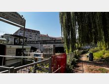 Ein kleiner Hafenbereich in Berlin mit einem Anlegesteg und einem Boot im Vordergrund. Im Hintergrund ist eine Brücke aus Stein mit Balustraden zu sehen, dahinter Wohngebäude, sowie ein Weg unter einer großen Weide und ein Metallzaun am rechten Bildrand.