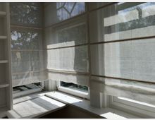 Ecke eines hellen Raumes mit großen, weißen Sprossenfenstern, die mit transparenten Raffrollos bedeckt sind. Im Hintergrund sind Bäume durch die Fenster zu erkennen.