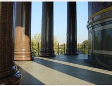 Blick durch mehrere massive Marmorsäulen auf eine Terrasse mit Steinboden, im Hintergrund sind Bäume und ein klarer Himmel sichtbar. Ein Geländer begrenzt die Terrasse.