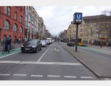 Straßenszene in einer Stadt mit mehrspuriger Fahrbahn, auf der Autos und ein Bus fahren, sowie einem separaten Fahrradweg. Links befinden sich rote Backsteingebäude, rechts stehen helle Wohn- und Geschäftshäuser. Ein U-Bahn-Schild mit der Markierung "U9" ragt rechts ins Bild.
