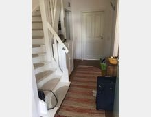 Heller Flur in einem Wohnhaus mit weiß gestrichener Treppe, Holzboden und einem rot-gemusterten Läufer. Rechts ein weißes Türblatt, links ein schmaler Schrank, daneben eine blaue Reisetasche und ein Korb mit Haushaltsgegenständen.