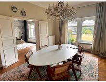 Esszimmer in einem eleganten Wohnraum mit Holzfußboden, weißem ovalem Tisch und Holzstühlen. Ein großer Kronleuchter hängt von der Decke, und durch ein großes Fenster mit Vorhängen fällt Tageslicht ein; im Hintergrund ein angrenzendes Zimmer mit gemusterter Tapete und Teppich.