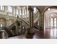 Großes historisches Treppenhaus mit elegant geschwungenen Marmortreppen, verzierten Geländern und hohen Rundbögen, umgeben von großen Fenstern, die Tageslicht einlassen. Die Architektur weist dekorative Elemente und Säulen im Neorenaissancestil auf.