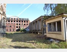 Ehemaliges Industriegelände mit überwuchertem Gras und mehreren verlassenen Gebäuden. Im Hintergrund ein mehrstöckiges Backsteingebäude mit zerstörten Fenstern, im Vordergrund ein kleines, verfallenes Häuschen mit zerbrochenen Scheiben.