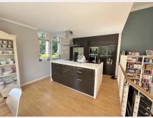 Moderne Wohnküche mit großer Kücheninsel, dunklen Hochglanzschränken und integrierten Backöfen, hellen Arbeitsflächen und Holzfußboden. Große Fenster mit Blick ins Grüne sowie angrenzende Regale und eine kleine Treppe auf der rechten Seite.