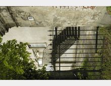 Fassade eines alten Gebäudes mit bröckelndem Putz, schwarzem Metallzaun im Vordergrund und einer steinernen Treppe, die zu einem Eingang führt. Im Bild sind außerdem ein Baum mit grünen Blättern und ein gepflasterter Weg sichtbar.