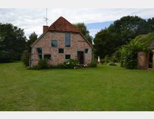 Fachwerkhaus mit roten Ziegeln und dunkelgrauen Holzbalken, umgeben von einem großen, gepflegten Garten mit Rasen. Im Hintergrund stehen Bäume, und rechts ist ein Nebengebäude sowie eine offene Holztür erkennbar.