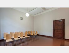 Ein schlichter Raum mit einer Sitzreihe aus hellen Holzstühlen, einer Wand mit einer analogen Uhr, moderner Deckenbeleuchtung und einer großen Holztür. Der Fußboden ist braun, die Wände sind beige gestrichen.