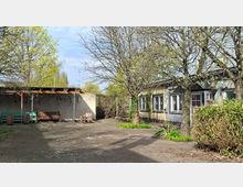 Gepflasterter Innenhof mit mehreren Laubbäumen und Sträuchern, rechts ein flacher Gebäudetrakt mit Fenstern und einer Tür. Links steht ein überdachter Sitzbereich mit Holzbänken vor einer Mauer.