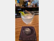 Ein Cocktailglas mit klarem Getränk und einer Limettenschale als Garnitur steht auf einer dunkelblauen Stoffserviette auf einer hölzernen Bartheke; im Hintergrund sind verschwommene Barutensilien und Flaschen zu erkennen.