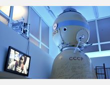 Raumkapsel mit der Aufschrift "CCCP" ausgestellt in einem Innenraum mit blauer Beleuchtung; an der Wand befindet sich ein Monitor, der das Bild eines Astronauten zeigt. Die Kapsel ist mit Unterschriften versehen und von technischen Details wie Kabeln und Metallkomponenten umgeben.