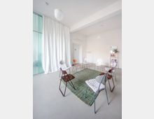 Heller, minimalistischer Innenraum mit grauem Boden und weißen Wänden, in dessen Mitte mehrere Stühle im Kreis um einen grünen Teppich stehen. Links hängt ein bodenlanger weißer Vorhang vor großen Fensterflächen, rechts sind eine Tür, eine Garderobe und ein Regal mit kleinen Gegenständen zu sehen.