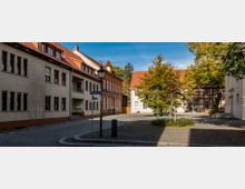 Kleiner Platz in einer historischen Stadt mit gepflastertem Boden, umgeben von Gebäuden im Fachwerk- und Backsteinstil sowie mit roten Ziegeldächern. Im Hintergrund stehen Bäume, und Straßenlaternen säumen den Platz.