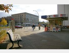 Straßenszene mit einem kleinen Zeitungskiosk auf der rechten Seite, einer Bank auf der linken Seite und einem großen, rechteckigen Mehrgeschossgebäude im Hintergrund. Menschen gehen auf dem Gehweg, während Autos an einer Kreuzung mit Ampeln vorbeifahren.