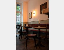 Gemütliches Café mit hölzernen Tischen und Stühlen, einer gepolsterten Sitzbank entlang der Wand und gedämpfter Beleuchtung durch eine Wandlampe. Im Hintergrund ist eine große Fensterfront mit Blick auf die Straße zu sehen.