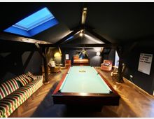 Ein stilvoll eingerichteter Dachraum mit dunklen Wänden, freiliegenden Holzbalken und einem zentralen Billardtisch mit grünem Filz. Im Hintergrund stehen gemütliche Sessel, ein Sofa mit buntem Streifenmuster, ein Klavier und verschiedene Steh- und Tischlampen.