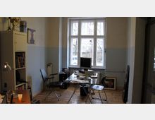 Ein schlicht eingerichtetes Büro mit Holzboden, einem großen Fenster mit Blick nach draußen, einem Glastisch mit Computer und zwei schwarzen Stühlen. Links stehen ein Bücherregal und eine Lampe, im Hintergrund sind Bilder und weitere Gegenstände an der Wand angelehnt.