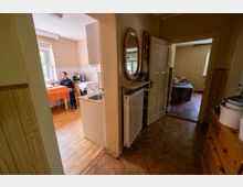 Blick durch einen Flur in eine ältere Wohnung: links eine kleine Küche mit gemusterter Tapete, Fenster und Esstisch, rechts ein schmaler Gang mit Holzfußboden, Rundspiegel und Heizkörper. Im Hintergrund führt eine offene Tür in ein Schlafzimmer mit Bett und gestreifter Tapete.