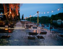 Ein Außenbereich eines Biergartens am Ufer eines Flusses, mit Holztischen und Metallstühlen, die auf Sand stehen. Lichterketten sind zwischen Holzpfosten aufgehängt, und Bäume sowie ein bewaldetes Flussufer sind im Hintergrund zu sehen.