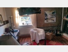 Wohnzimmer mit Holzboden und einem großen Fenster, das von hellen Vorhängen umrahmt ist. Ein weißer Sessel steht vor einer rustikalen Steinwand mit einem Landschaftsbild, daneben eine Bücheransammlung und eine Gitarre; ein Schreibtisch mit Lampen und Büchern ist links zu sehen.