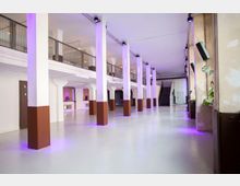 Industrieartiger Veranstaltungsraum mit weißen Säulen, die bis zur Decke reichen und an der Unterseite braun lackiert sind. Im Hintergrund befinden sich eine Treppe sowie eine offene Galerie im ersten Stock mit Metallgeländern; lila Beleuchtung verleiht dem Raum Akzente.