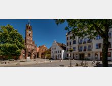 Historischer Platz mit einer gotischen Backsteinkirche mit Turm und Uhr im Vordergrund, flankiert von farbenfrohen, historischen Gebäuden und moderneren Fassaden; im Zentrum ein kleiner Brunnen, umgeben von Bäumen, Fahrrädern und Laternen.