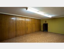 Ein leerer Raum mit gelblich gestrichenen Wänden, einem Fischgrät-Parkettboden und eingebauten Holzverkleidungen an einer Wand. An der rechten Seite befindet sich eine geöffnete Tür, und an der Decke sind lange, rechteckige Deckenleuchten befestigt.